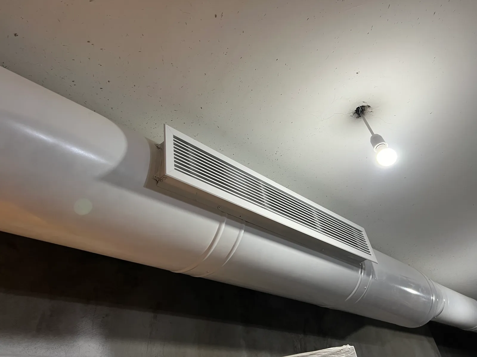 An exposed white round spiral duct fitted with a linear supply grille below the ceiling.