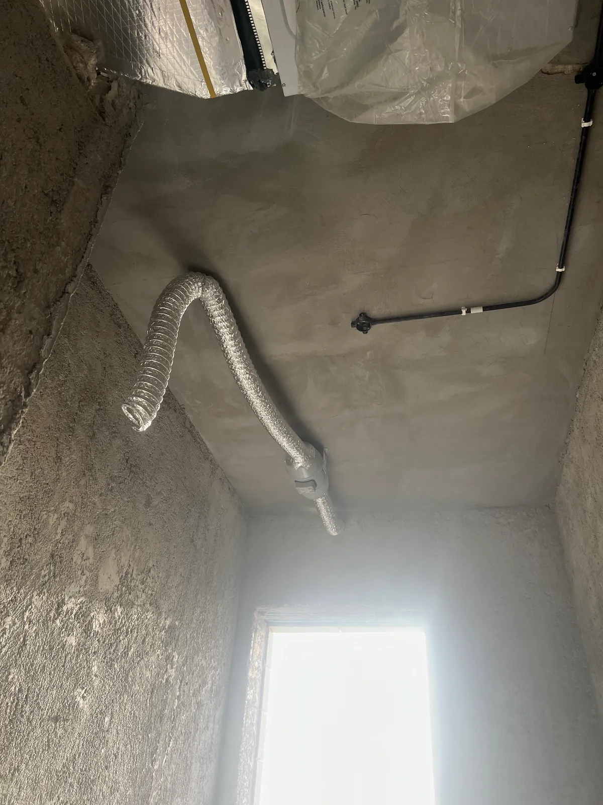 A flexible aluminium ventilation duct connected to an inline fan in a shaft during the construction stage.