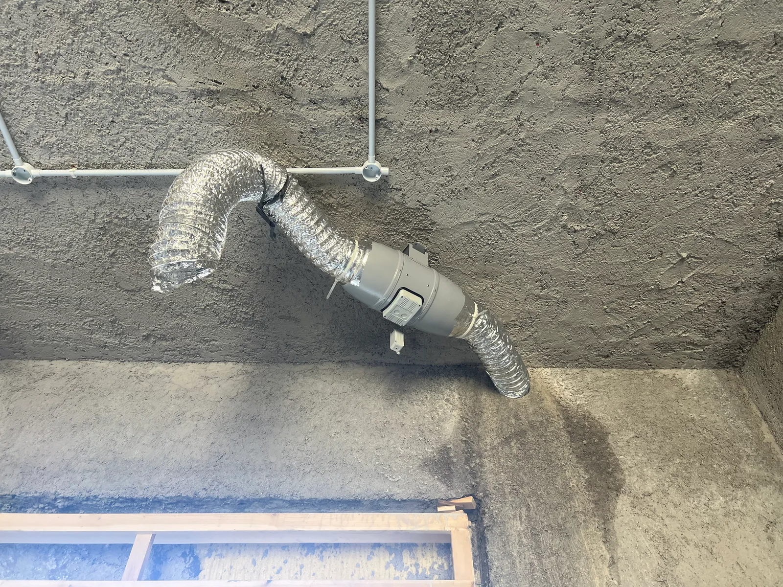 An inline ventilation booster fan connected to flexible ducts on a concrete wall during construction.